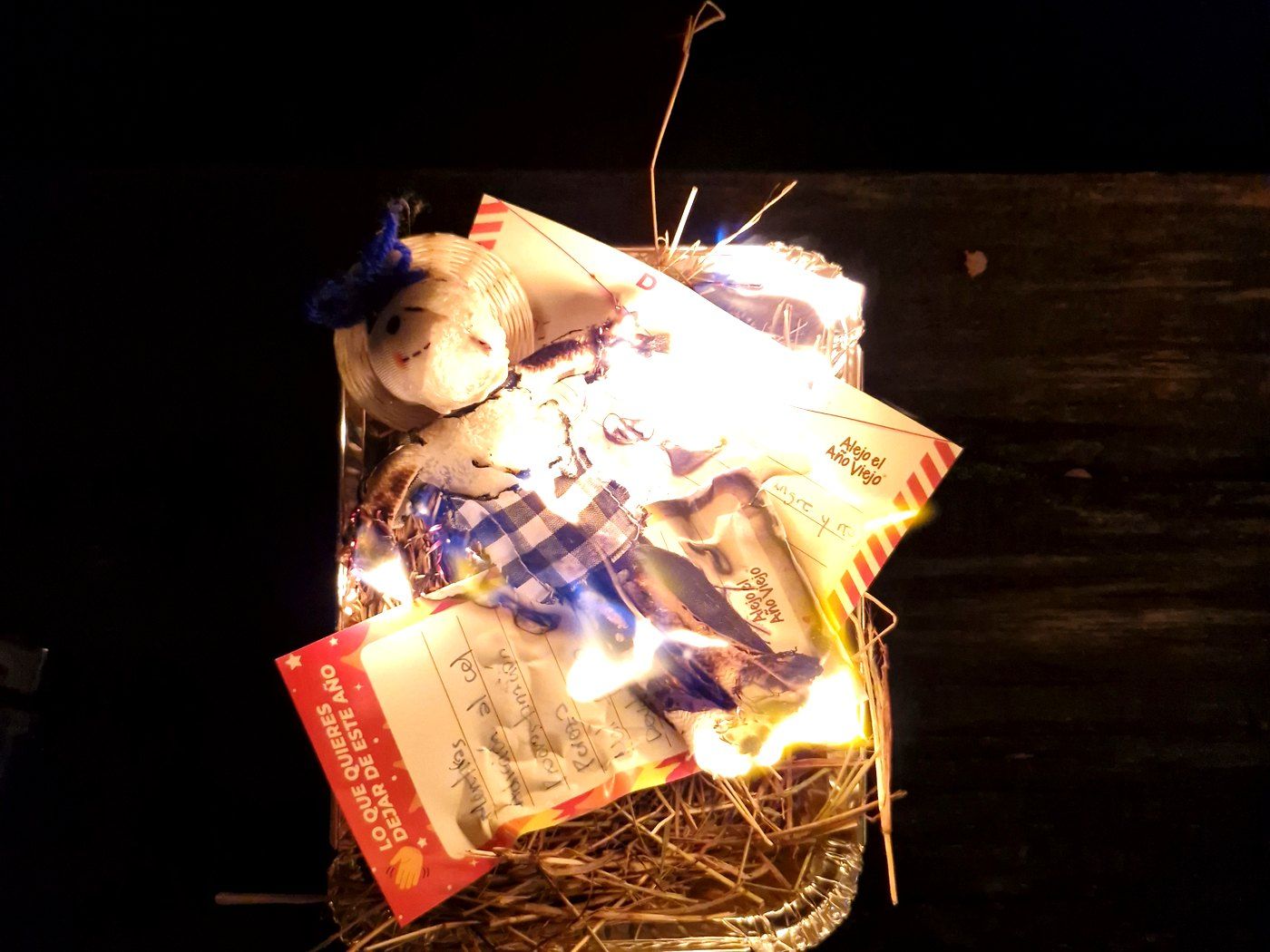 Muñeco de Año Viejo ardiendo de noche sobre un tejido de paja, con las llamas iluminando la tela y el papel del relleno.